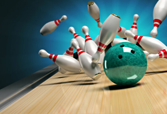 a green bowling ball hitting pins and the pins flying in different directions.