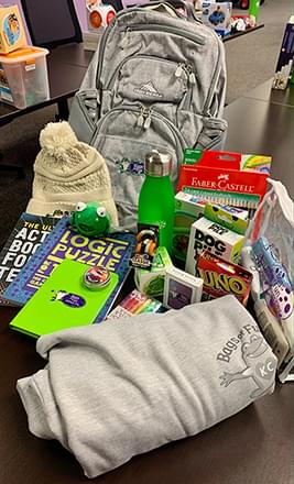 A gray bags of fun bag on the table with items laid out in front of it. These items include a winter hat, puzzle books, games like Uno, colored pencils, a water bottle, and a gray Bags of Fun shirt.