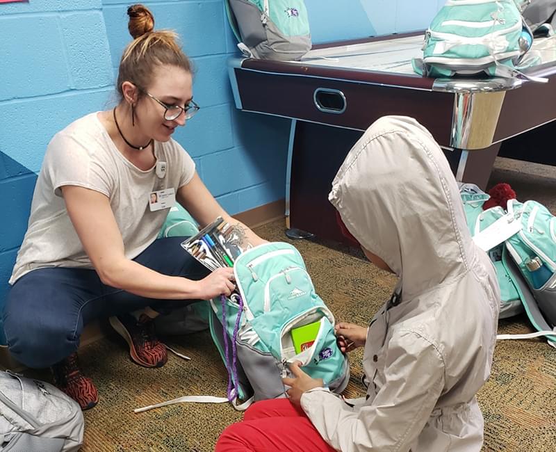 a volunteer is sitting with a child and there is a teal bag of fun between them. The volunteer is holding some markers that are half in and half out of the bag (backpack). The child is opening or closing the front pocket of the backpack.