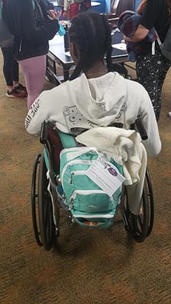 The back of a wheelchair with a teal bag of fun hanging on it. There is a teenager sitting in the wheelchair.