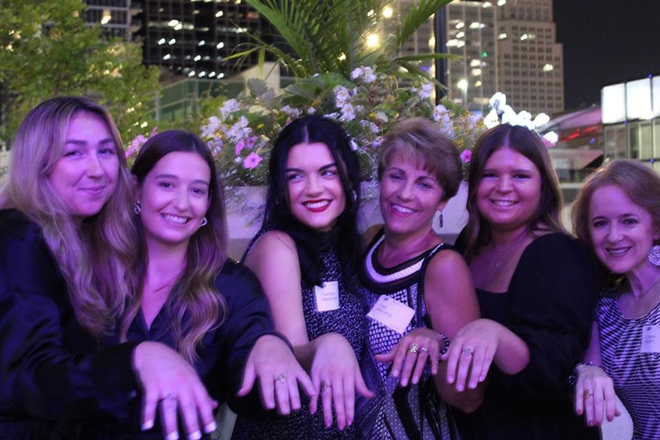 6 women smiling for the camera with a hand out showing off their ring from the event. They are outside in the evening time and there are buildings with lights in the distance. They are standing in front of lots of plants with flowers.