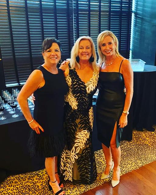 3 women, one of which is the owner of Bags of Fun, Tammy, standing in their black and white dresses and smiling for a picture. There are glasses of wine on a table behind them.