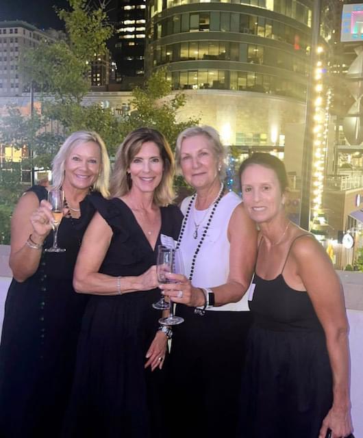 4 women in their black and white dresses holding champaign and smiling at the camera. They are outside with buildings with lights behind them.