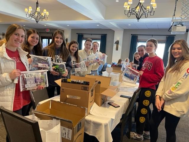 a group of 10 teenage girls standing around a long table with open amazon boxes. The girls are smiling at the camera and each holding a hygiene kit that will get packed into a Bag of Fun.