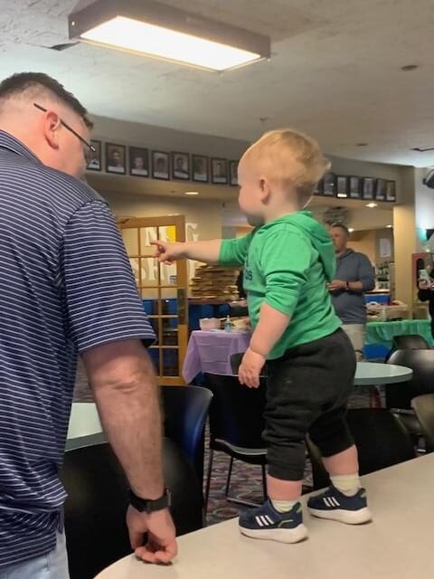 A toddler wearing a green shirt standing on a table pointing to something. His Dad is standing next to him and looking at him. There backs are towards us.