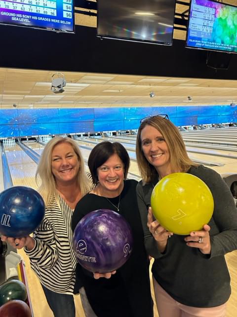 3 women holding their bowling balls up and smiling at the camera. One bowling ball is yellow, one is blue, and one is purple. You can see the bowling lanes behind them.