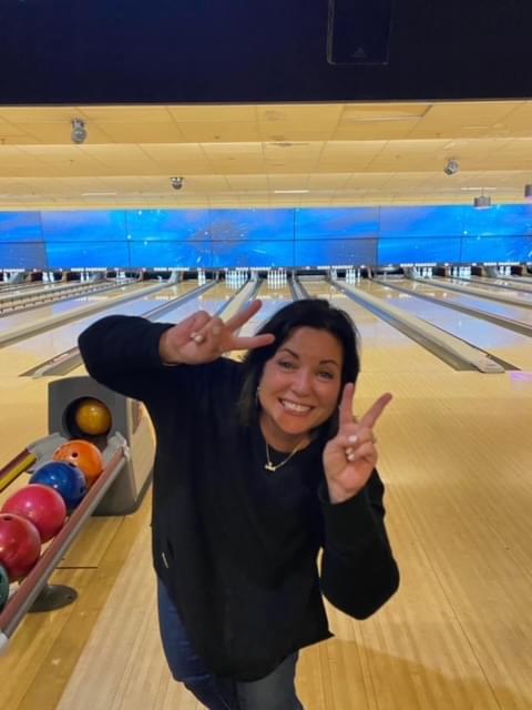 a woman smiling at the camera with both hands up showing the piece sign to the camera. You can see the bowling lanes behind her.