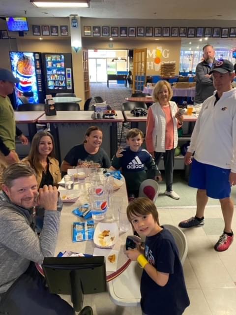 A group of adults and kids sitting at the table at their bowling lane. There is food on the table and they are smiling at the camera.