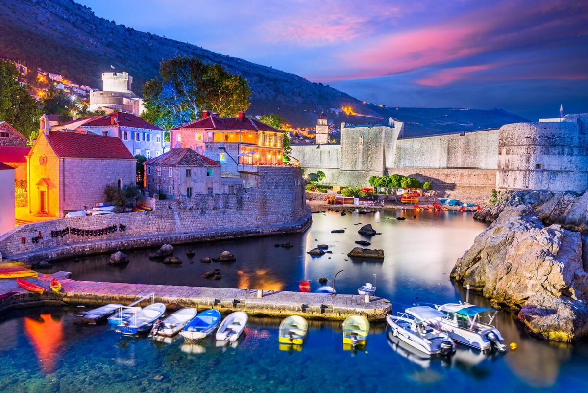 A stunning nighttime view of a town on the water. The sky is a dark shade of blue and purple with some reddish orange. There is a lake that goes through the city and a dock with boats. We see large mountains in the distance and the town seems to slope up from the water as the mountain slopes up.