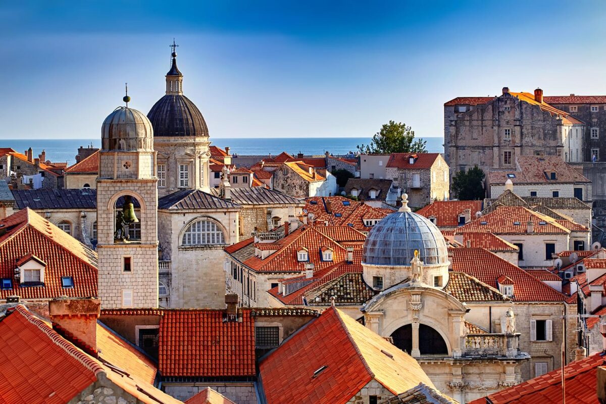 The tops of buildings in a compact city. Many of the buildings have red roofs and there are a couple with domes. We can see the ocean in the distance.