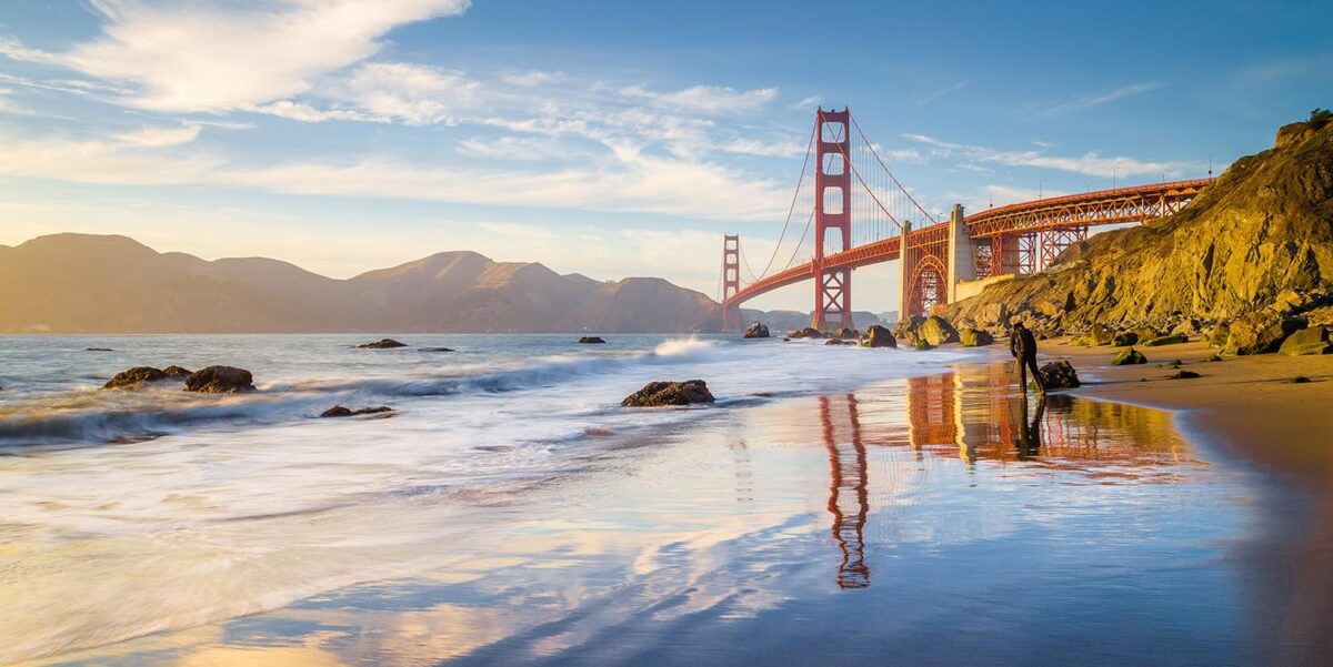 We are looking up at the Golden Gate Bridge from a beach with crashing waves. The sun is setting and there as the water glistens with the orange hues. There is a man walking close to the water.