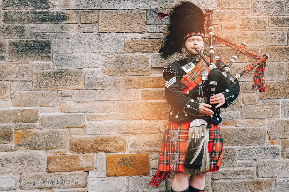 A Scottish man with the full Scottish kilt outfit playing the bagpipes. He is standing in front of a red and brown brick wall.