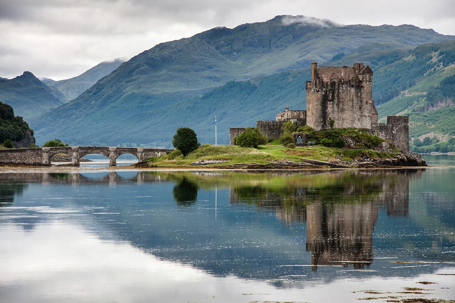 A view of a Scottish castle surrounded by water. There is a bridge that leads to the castle. There are also large hills in the background and some fog has set in at the tops of the hills.