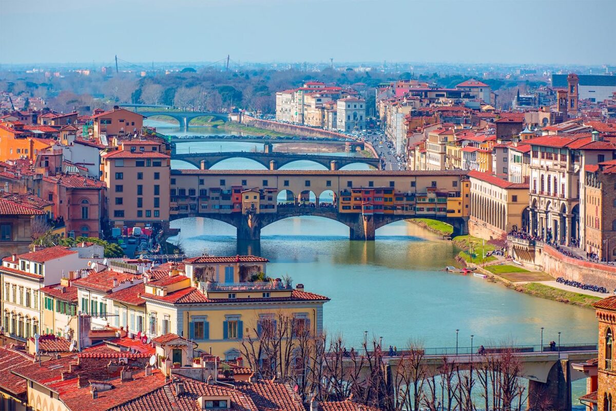 a city with lots of older looking Italian buildings with red roofs. There is a lake that runs through the city and four bridges going from one side to the other.