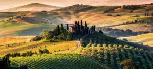 A view of the rolling hills of the Tuscany countryside. There is an orange glow as the sun shines on parts of the hills. You see small roads and one large house or winery at the top of a hill with trees around it.