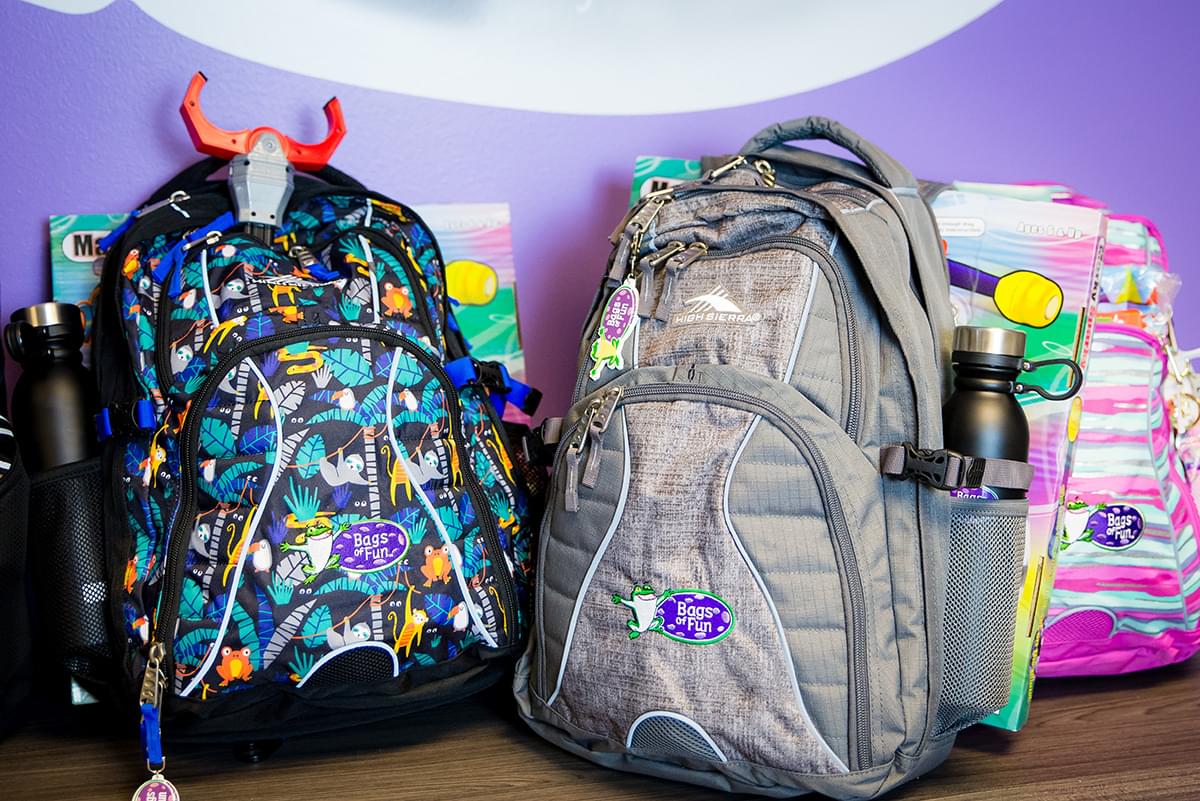 There are two backpacks in the foreground. One is black with illustrations of sloths, tropical palm trees, birds, snakes, frogs and other animals on it. There is a black water bottle in the side bottle holder and a toy red, black, and gray grapple claw sticking out of the top of the large main pocket. There is also a gray backpack next to it but all the pockets are closed. It also has a black water bottle in the side bottle holder. In the background there is a pink and light blue unevenly striped backpack. The backpacks are on a brown wood table with a purple wall behind them.