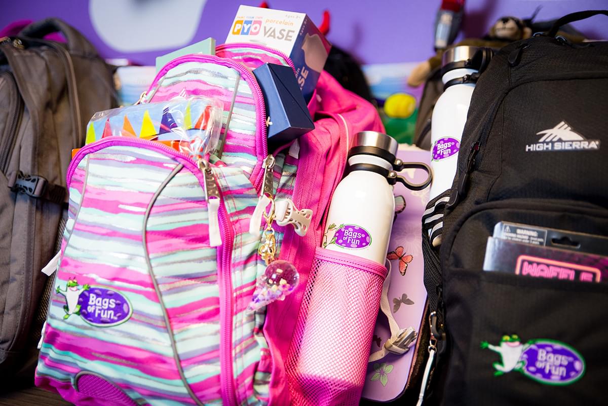 A close up image of the pink, light pink, blue, and gray unevenly striped backpack. The pockets are open and we see a PYO Vase coming out of the top of the large pocket. There are toys coming out of the two smaller pockets but it is hard to make out what they are. There is a white Bags of Fun water bottle in the side bottle holder. There is also a solid black backpack next to the pink one. We can only see half of it but there is a toy box in the front small pocket and a white Bags of Fun water bottle on the side. In the back we also see part of a sold gray backpack and the purple wall behind the backpacks.