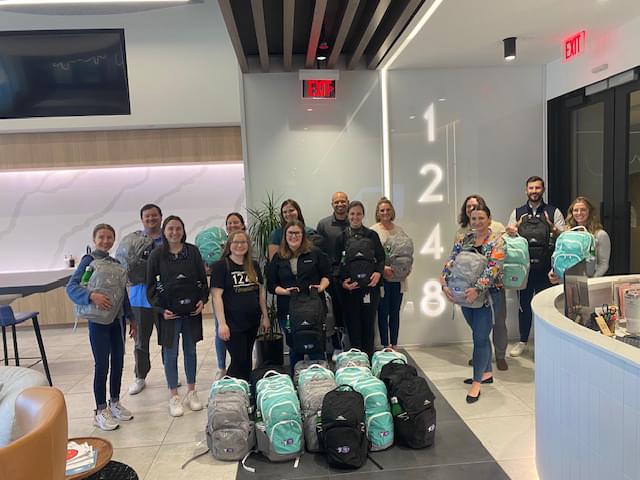 1248 team posing for a picture in their office. They are holding bags and there are bags on the floor in front of them The office has tall ceilings and if very modern looking with wall lights, white walls, and the number 1248 projected on the wall.