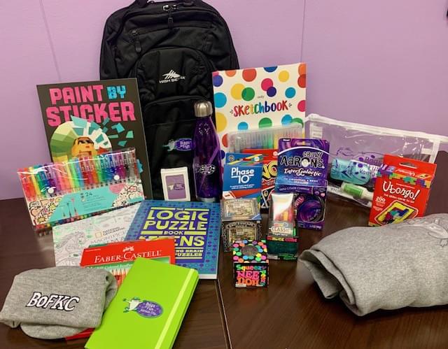 All of the items in a bag are out on a table with a black backpack behind. There are a pack of colored markers, sketchbook, Paint By Stickers, toys, a blanket, and a KC beanie hat.