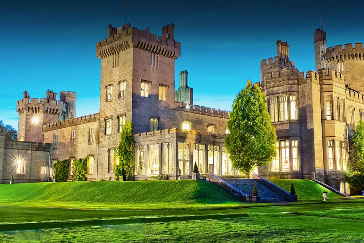 We see a large multilevel light brown brick castle. With glimmers of bright light in the windows. The castle sits on a small grassy hill, just a couple feet up. there are stairs that lead down to more open grass. The lights and sky suggest it is evening time but there is still some blue light in the sky.