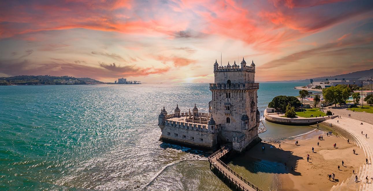 a panorama style photo of a small castle along the water with a beach behind it. The castle is just one tower a square open top building in front of it. There is a beautiful red and muted orange sky behind.