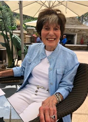 Ann Solomon sitting in a black chair outside with a large restaurant umbrella behind her. She is smiling and wearing white pants, a white shirt, and a blue denim looking jacket that has one button buttoned.