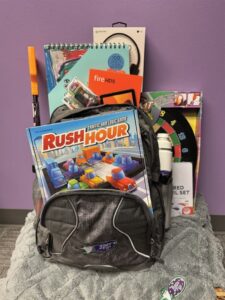 A backpack with Rush Hour traffic jam logic game in the front and a Bags of Fun water bottle in the side cup holder, and a Amazon Fire, art pad and headphones in the back. The Bags of Fun backpack is sitting on a Bags if Fun blanket, and it is resting on a packaged dart board.