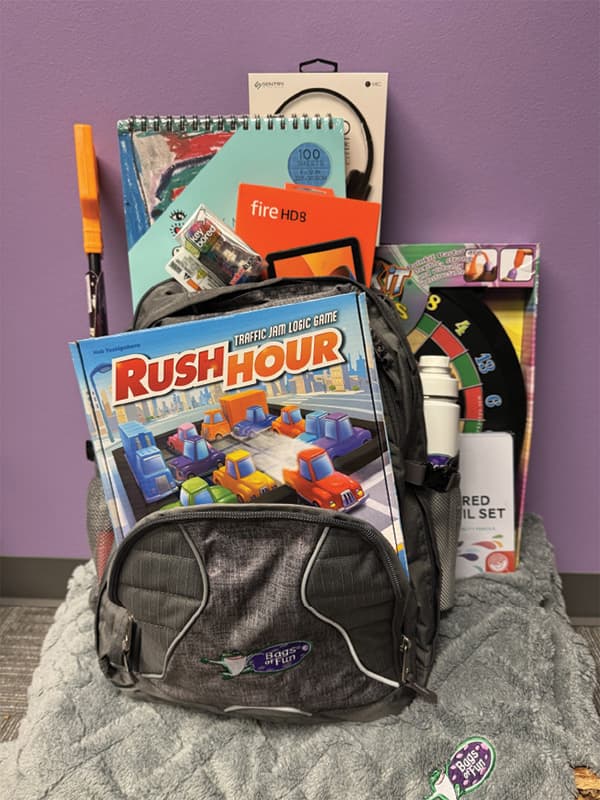 A backpack with Rush Hour traffic jam logic game in the front and a Bags of Fun water bottle in the side cup holder, and a Amazon Fire, art pad and headphones in the back. The Bags of Fun backpack is sitting on a Bags if Fun blanket, and it is resting on a packaged dart board.