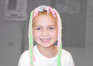 Gabby smiling at us. She has these fun and silly green, pink, and blue strings of yarn covering the top of her head and hanging down both sides of her head. She has such a genuine happy smile on her face. The background is grayed out but she is in full color.