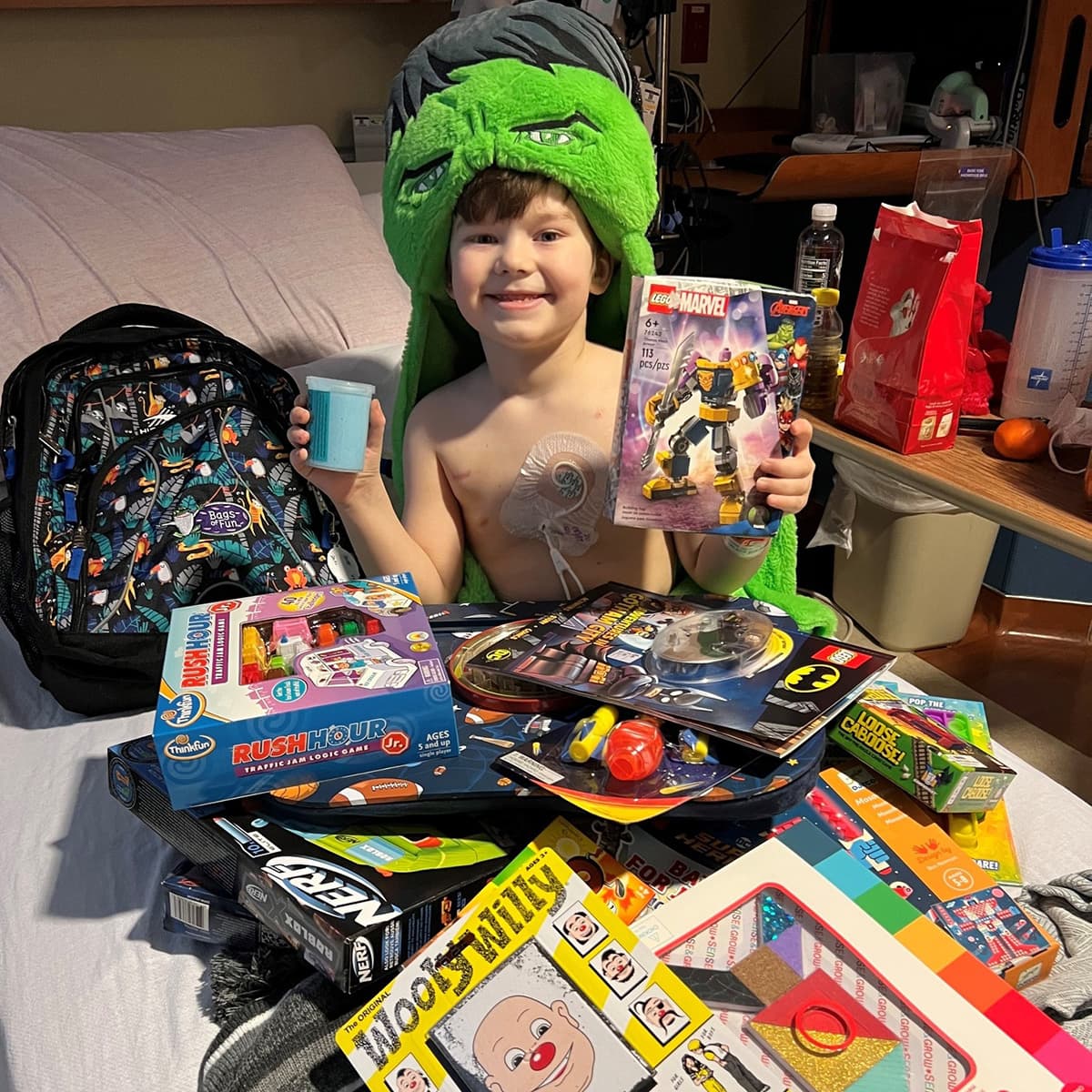 Kid with a green fuzzy Hulk hat holding some toys and having lots of toys all around him as he sits in his hospital bed.
