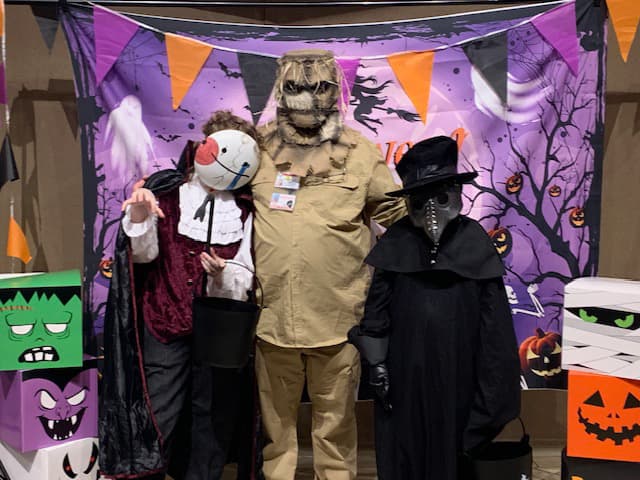 3 people in very creepy costumes posing in front of a Halloween tree and witch scene backdrop.
