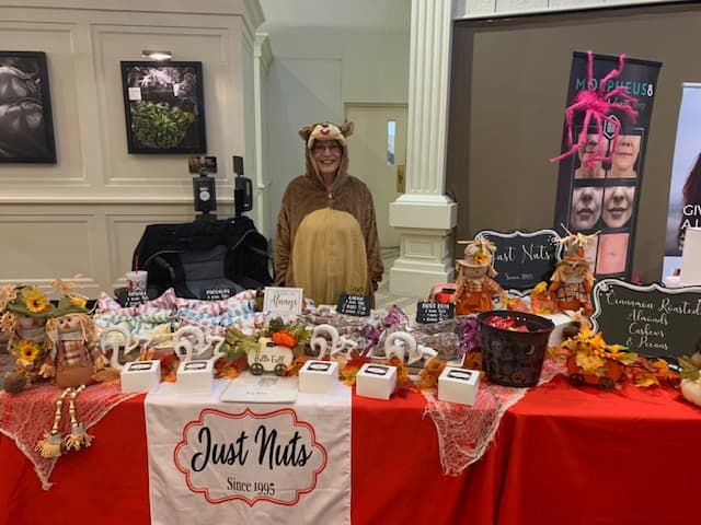 Someone in what appears to be a full squirrel or reindeer onesie behind the Just Nuts table, selling roasted almonds, cashews, and pecans.