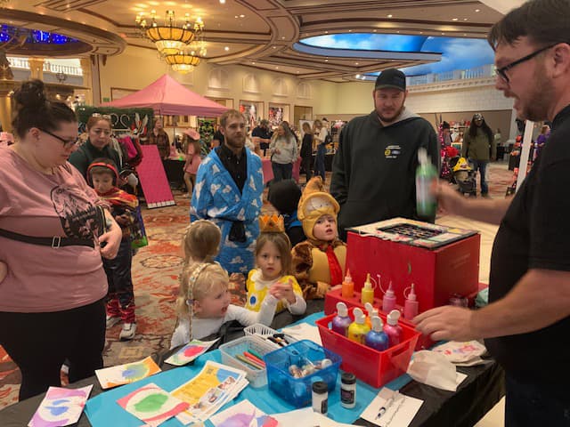 Kids and parents at an art station where someone is holding paint and creating splatter or spiral art with a machine for each kid.