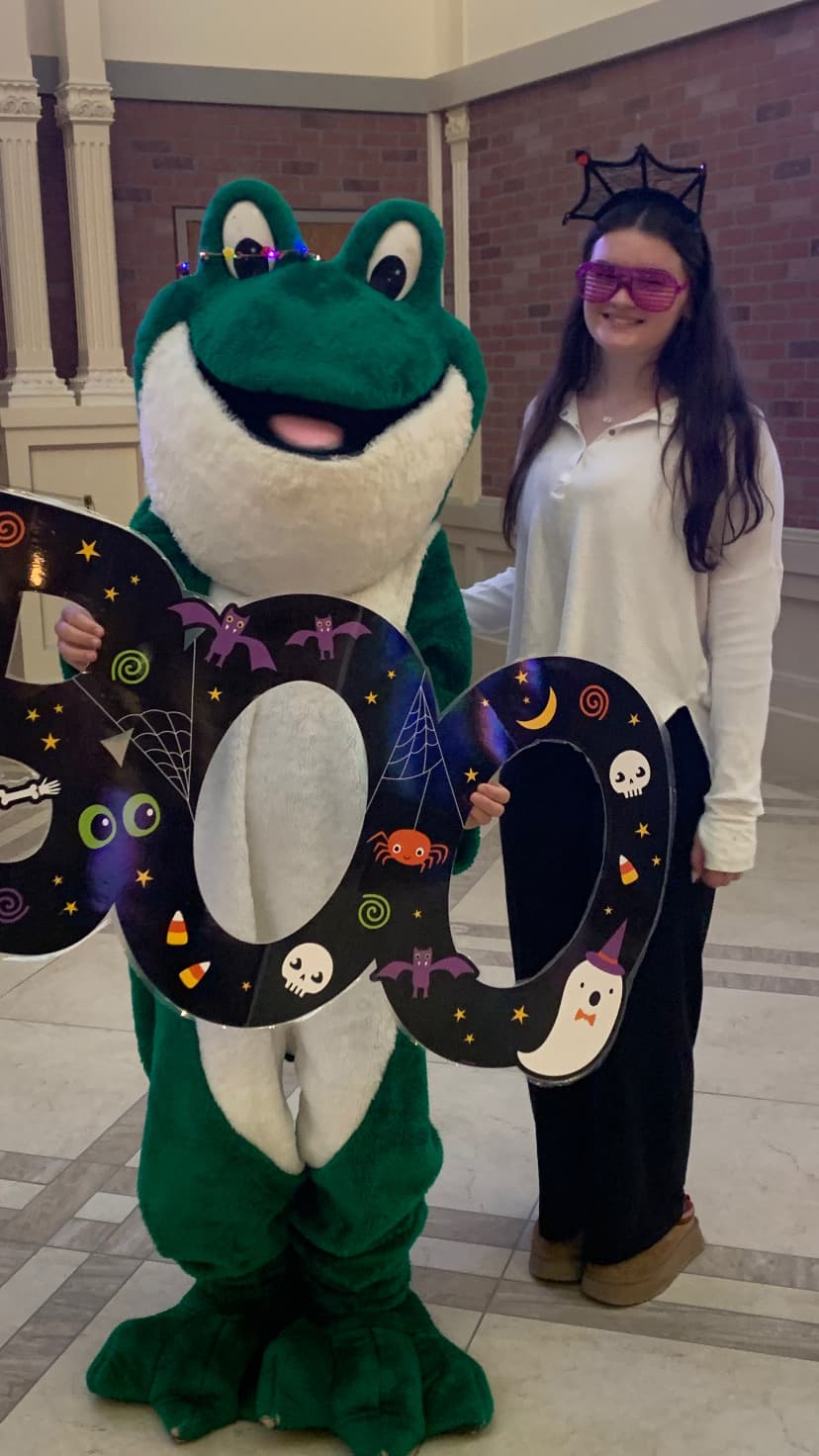 Someone standing with the Bags of Fun Frog mascot, who is holding a sign with the word Boo cutout. The sign is black with halloween illustrations.