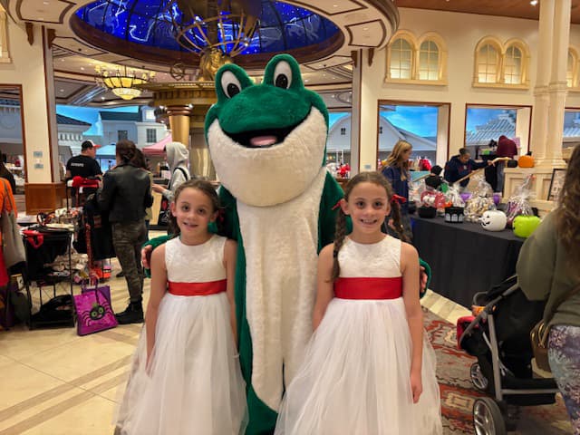 The Bags of Fun frog mascot posing with two girls, that look like twins, in white dresses with red ribbon belts.